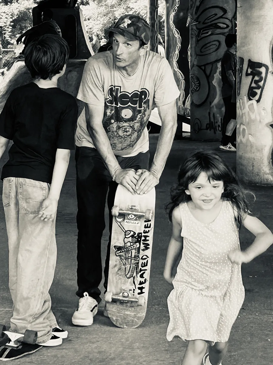Black and white photograph of Duane Pitre talking with young skaters at a skatepark, holding a skateboard